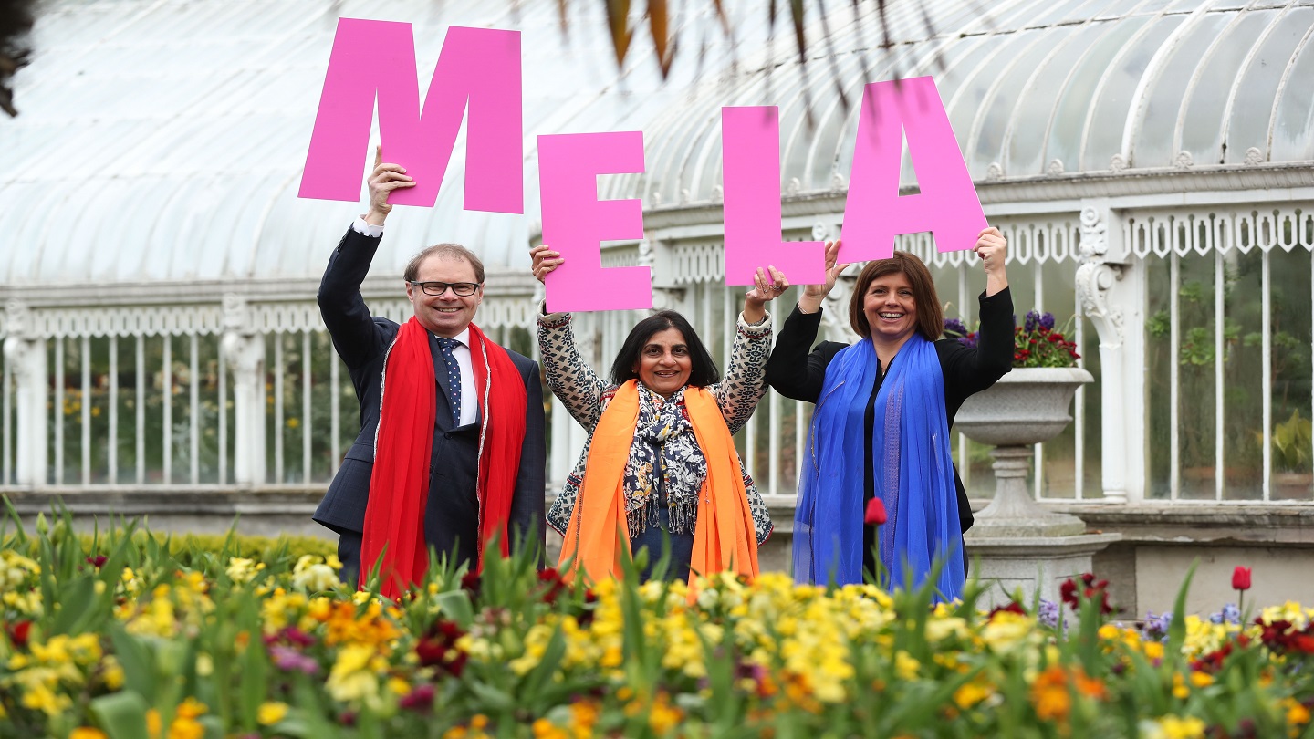Three people stand outside in a botanical garden. They're wearing colourful scarves and holding up large pink letters that spell 'MELA'