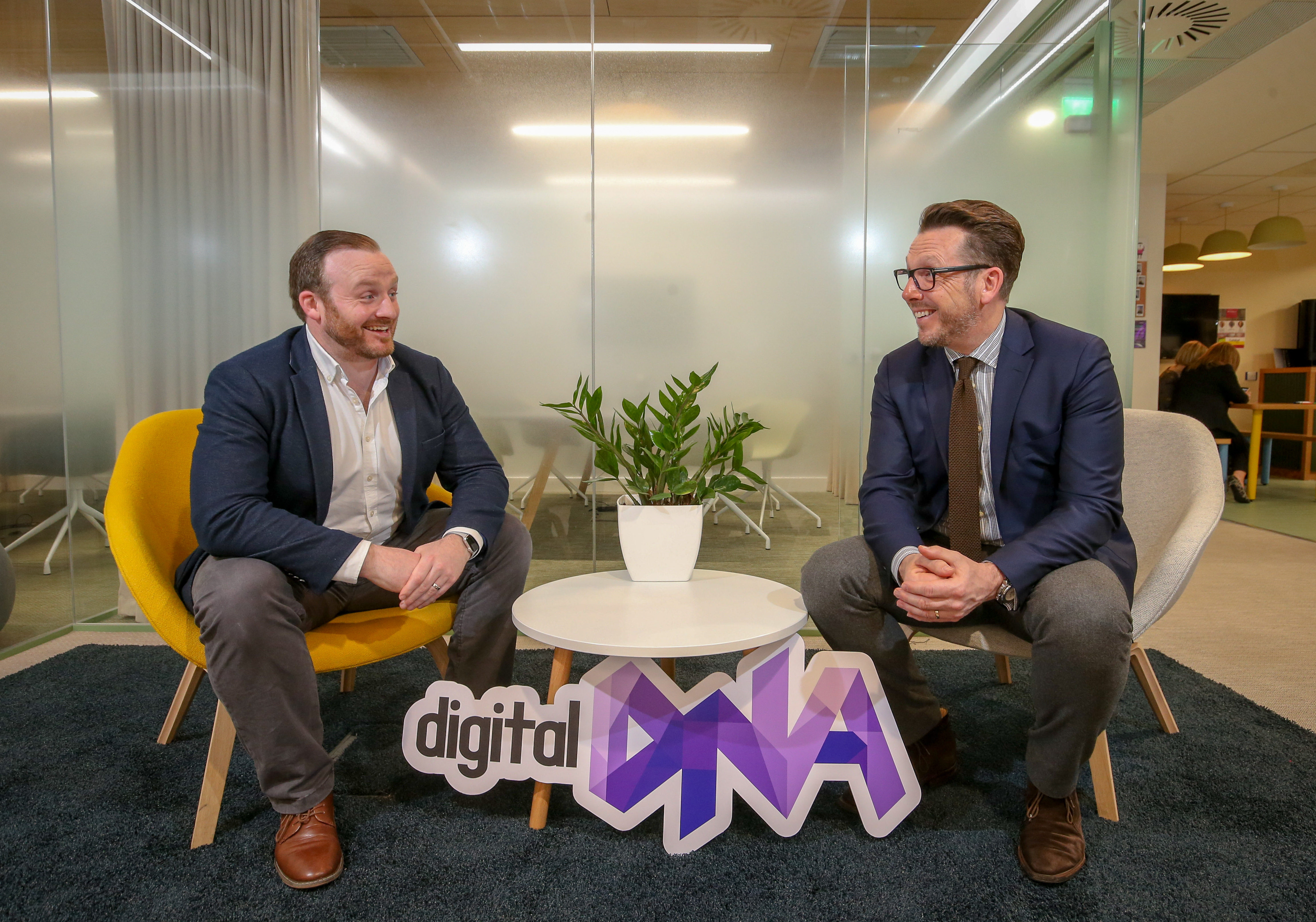 Two men in suits sit opposite each other at a coffee table, looking at each other and laughing. a purple 'DIgital DNA' sign sits in front of them.