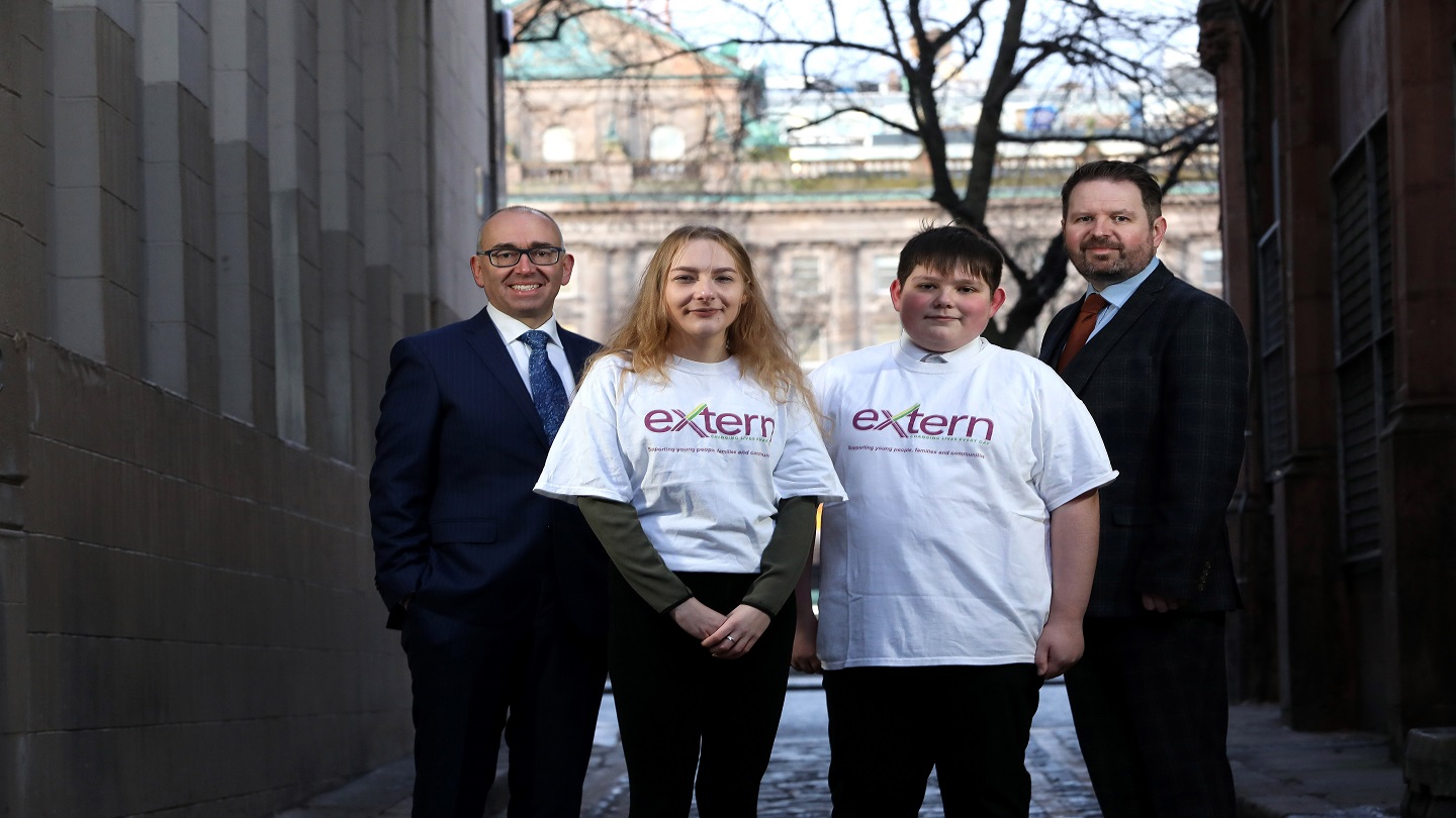 Two business men stand at either side of a young woman and man, who are both wearing white t-shirts that say 'extern'. They are standing outside, in between two buildings with Belfast City Hall behind them.