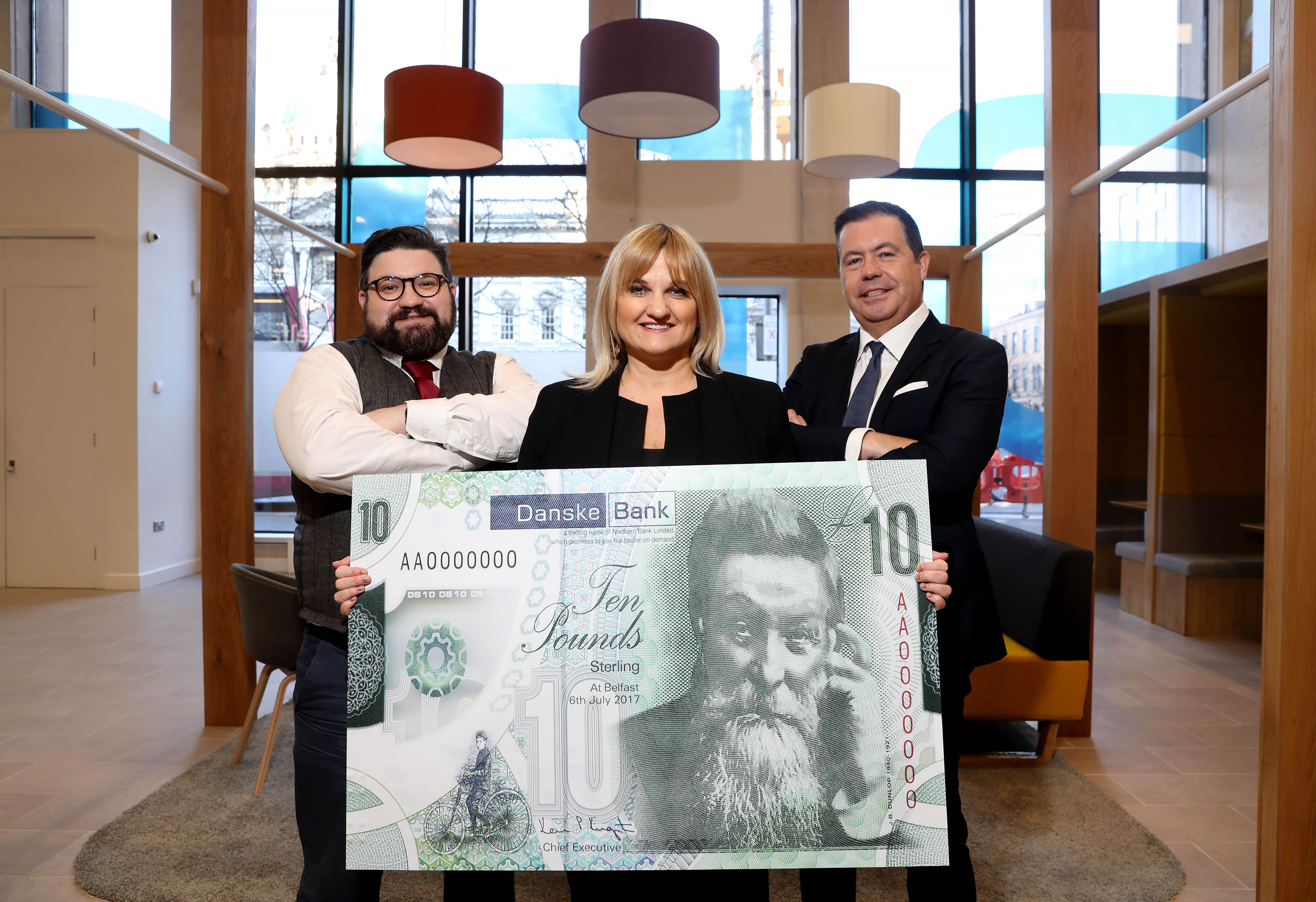 A woman stands between two men holding a large, green Danske Bank £10 note. The two men have their arms folded. 