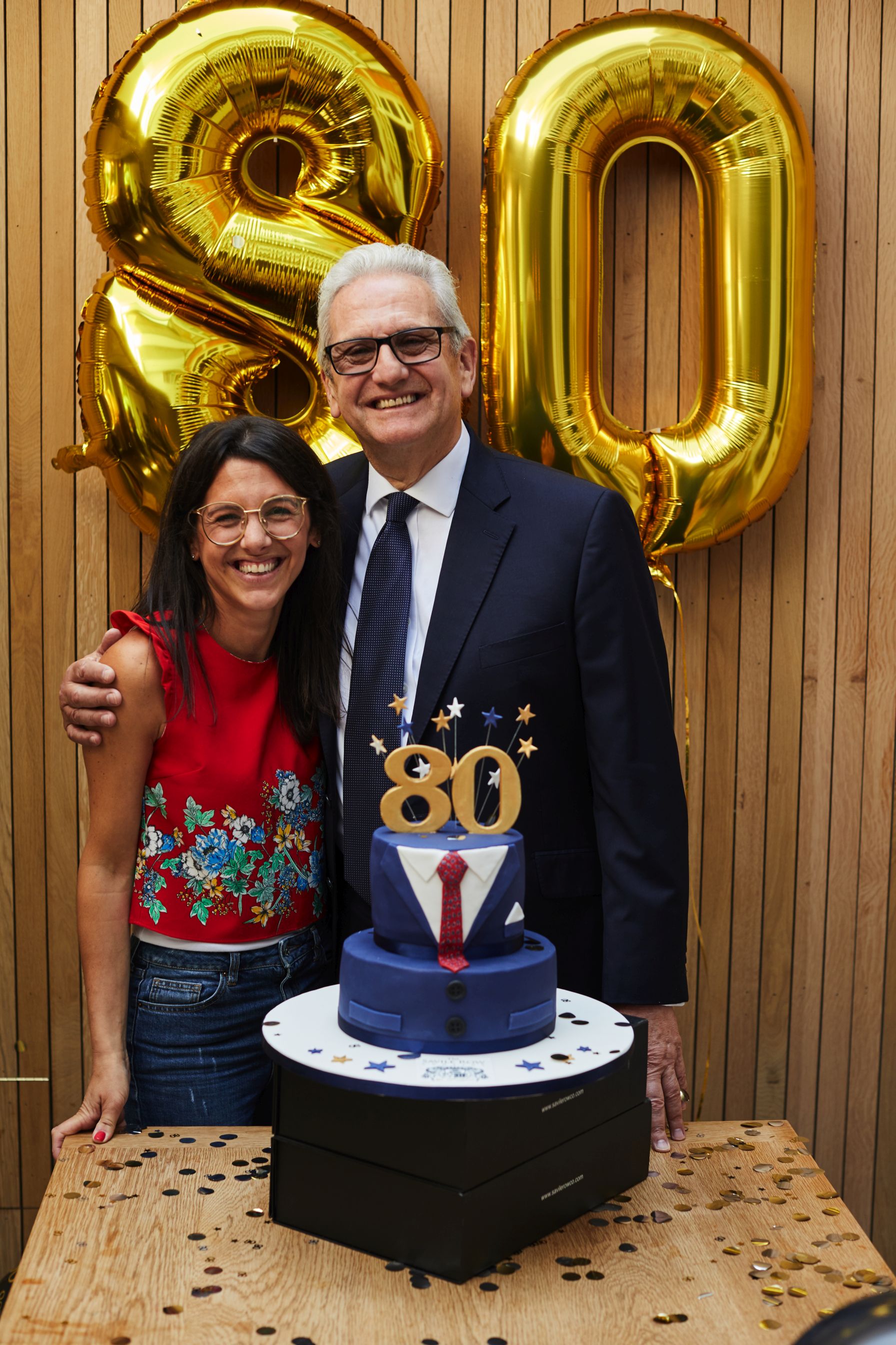 A woman and man stand together side by side smiling widely. There are two big '8' and '0' balloons behind them and a cake decorated like a suit with an '80' on top.