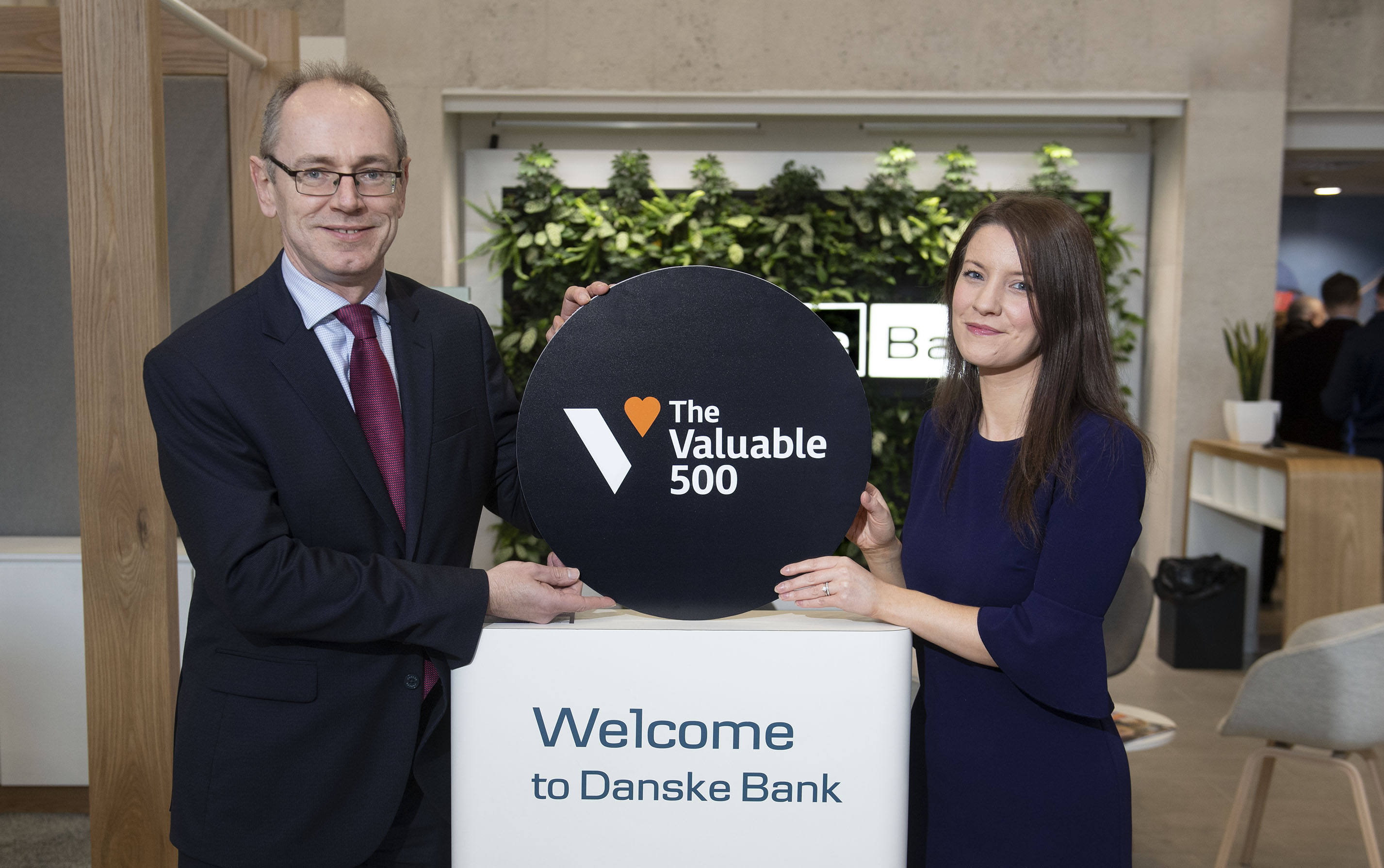 A man and woman stand inside. They're holding a circle board that says 'The Vaulable 500' above a stand that says 'Welcome to Danske Bank'.