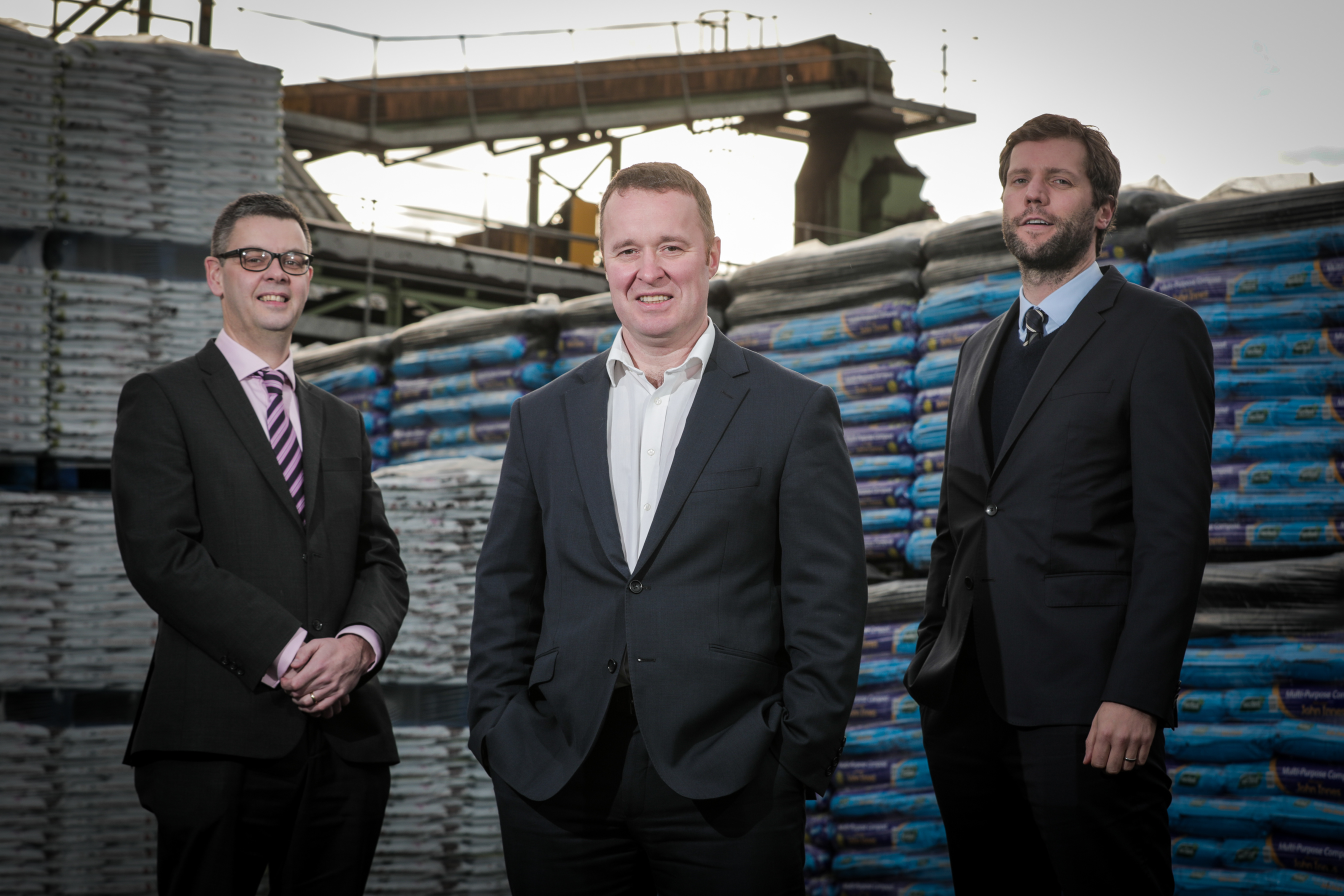 Three men in suits stand, spread out from each other slightly, in a workyard. Behind them is machinery for transporting products and stacks of bags of soil.