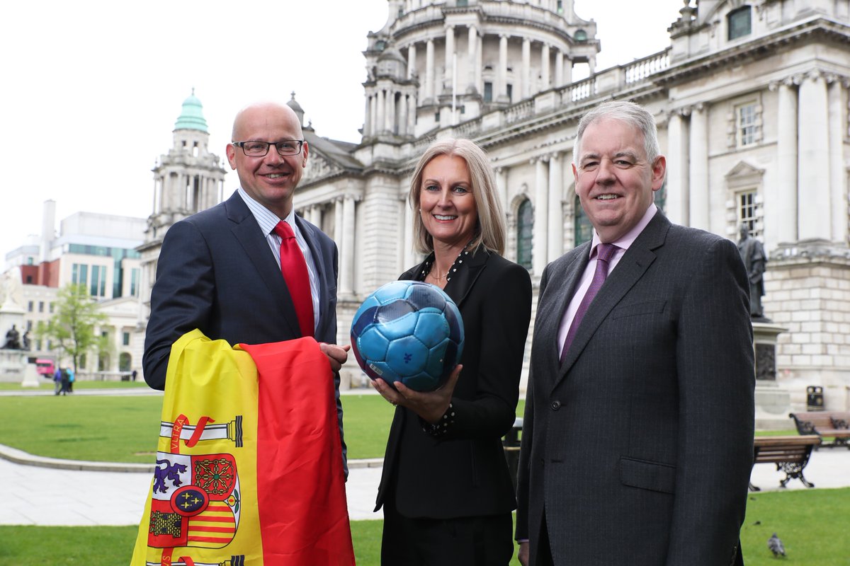 Three people, a woman in the middle holds a football and a man to her left holds a Spanish flag. They are outside, in front of the grand Belfast City Hall.