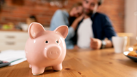 Budgeting blog - May 22 A pink savings 'piggy bank' sits in focus on a table, behind and out of focus is a couple looking towards the piggy bank.
