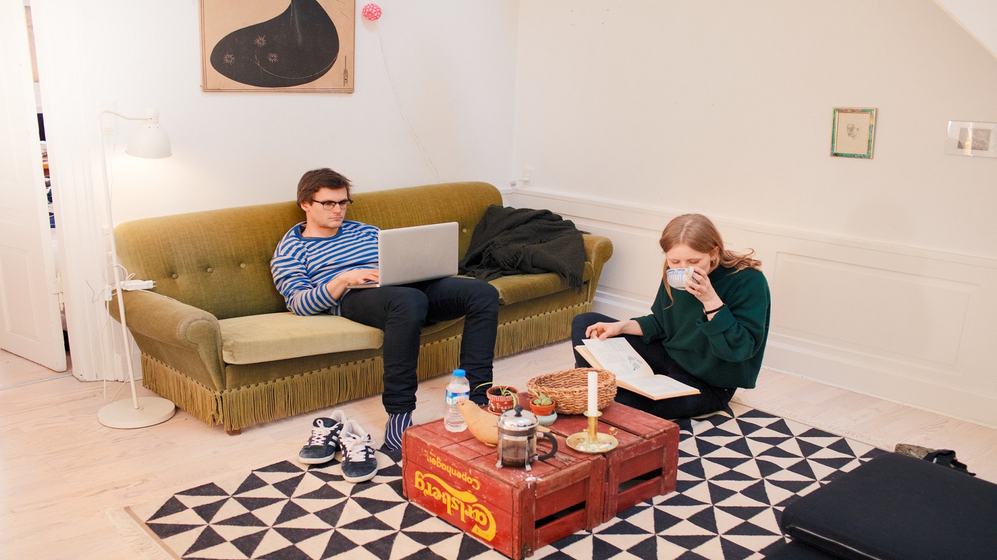 Two young people sit in a living room - one is lounging on a sofa with a laptop on their knee and the other is cross-legged on the floor with a book and a mug of hot drink.