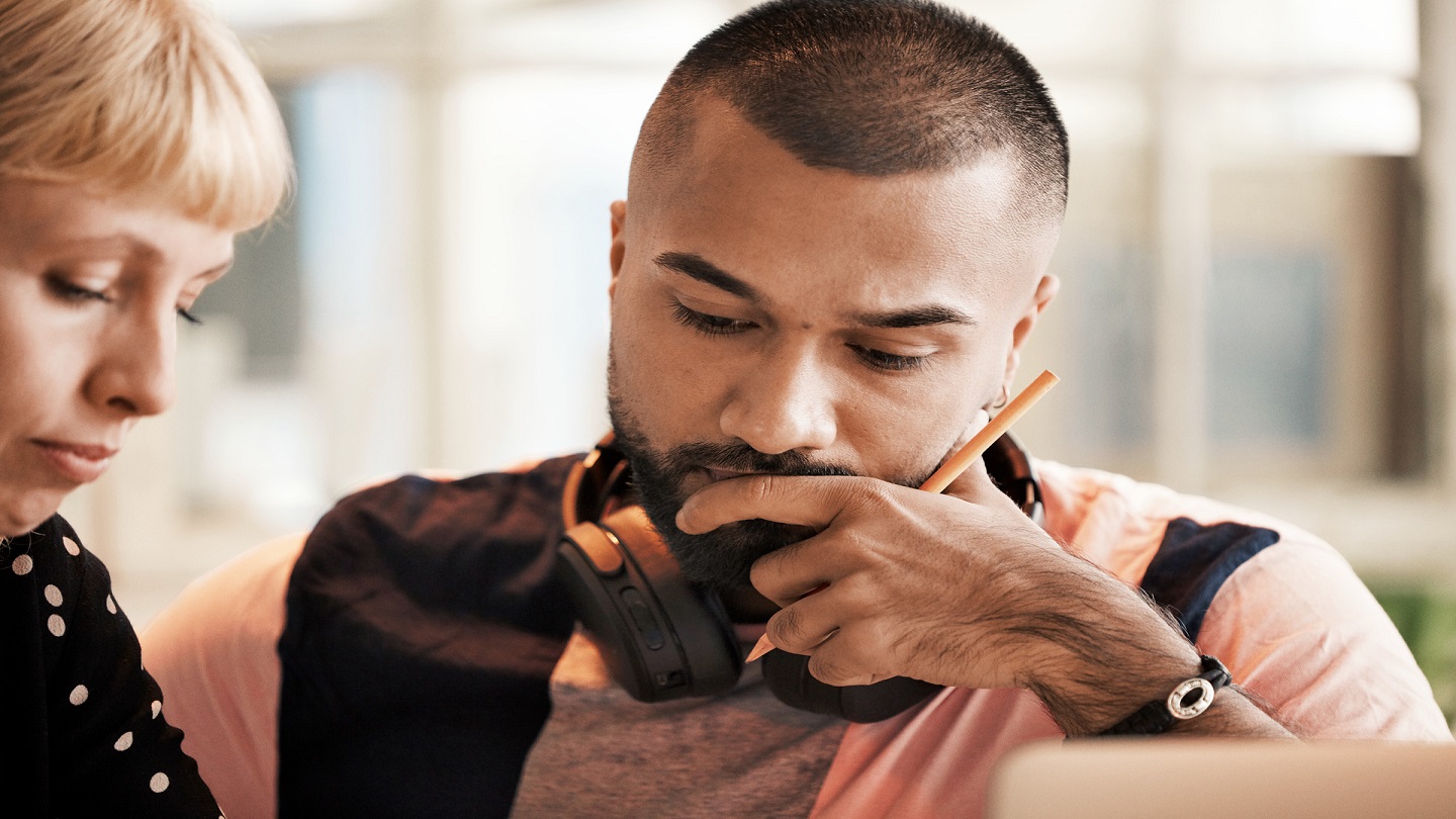 A man and woman sit close by each other. The woman is looking down at something, and the man has headphones round his neck, a pencil in his hand and a finger up to his lip. He's looking down at something thoughtfully - they are looking together.