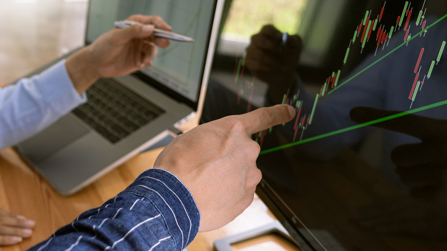 Two individuals analysing financial data on a computer screen displaying a candlestick chart, with one person pointing to the screen and the other holding a pen.
