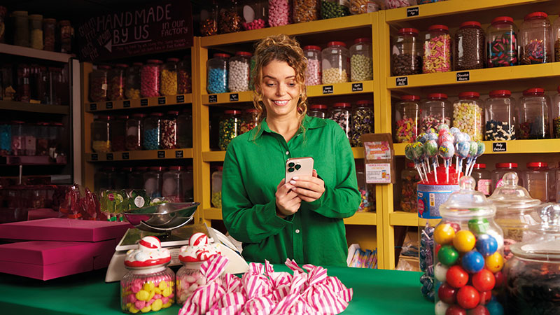 A person standing in a colourful sweet shop filled with jars of various candies, holding a smartphone. The counter displays jars of sweets, striped candy bags and lollipops.