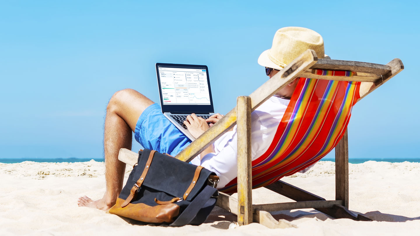 Person sitting in a striped deck chair on a sandy beach using a laptop, with a bag placed beside the chair and the sea in the background.