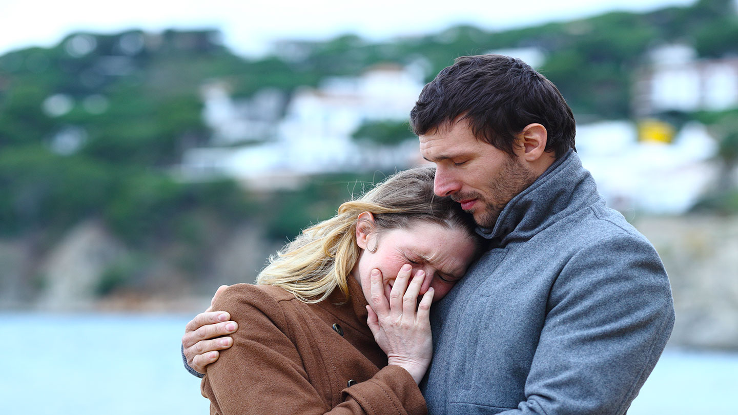 A couple stands outdoors near a waterfront, with the woman leaning her head on the man's chest. The man wears a grey coat and holds the woman close, offering comfort. The background features blurred greenery, cliffs, and buildings.