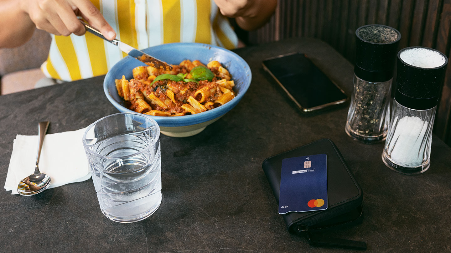 Person dining at a table with a bowl of pasta, a glass of water, salt and pepper shakers, a mobile phone, and a Danske Bank debit card placed on a wallet.