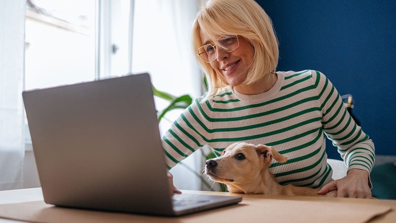 Woman using a laptop at a desk with a dog resting on her lap in a home environment.