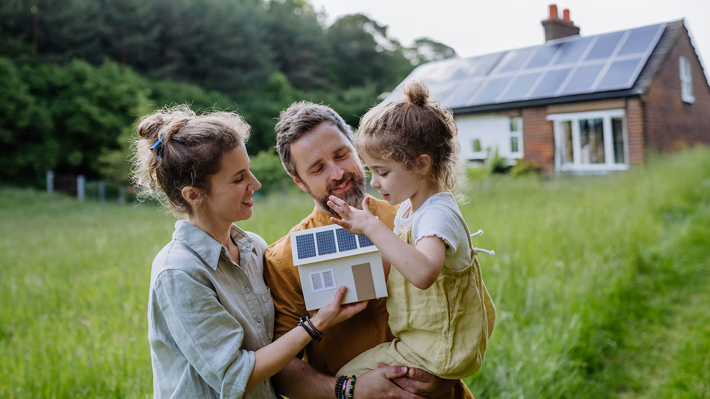 Family holding a small model of a house with solar panels in a grassy field, with a solar-powered brick house in the background.