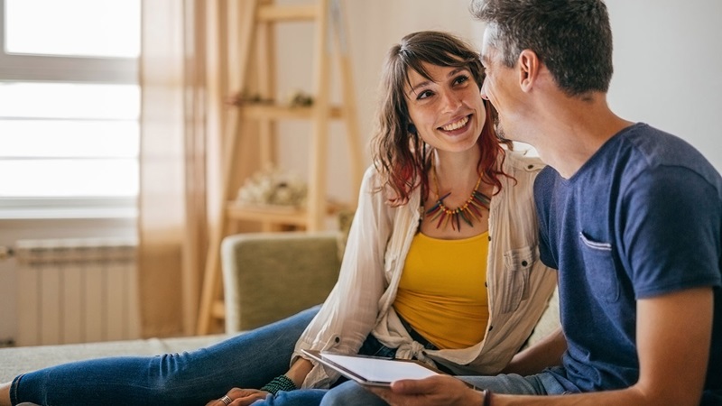 Happy couple sitting on a sofa in a cosy, well-lit living room, with one holding a tablet and the other wearing casual clothing and a colourful necklace.