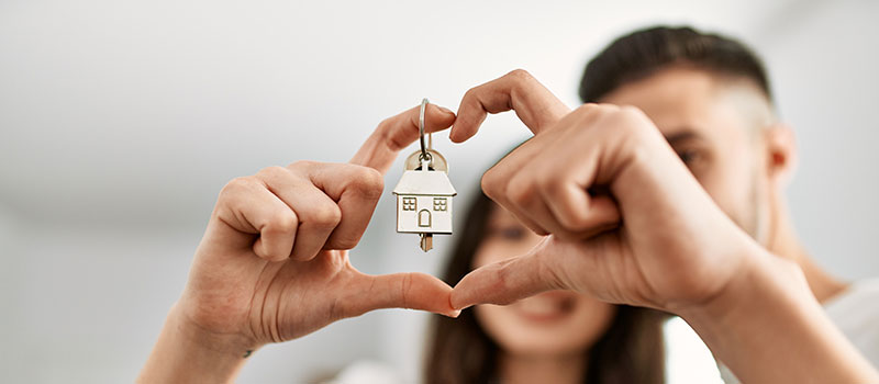 Close-up of hands forming a heart shape while holding a keychain with a house-shaped pendant, symbolising home ownership.