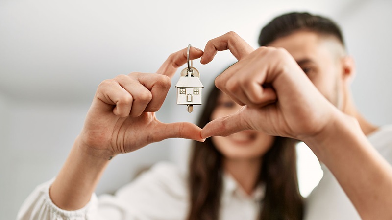 Close-up of hands forming a heart shape while holding a keychain with a house-shaped pendant, symbolising home ownership.