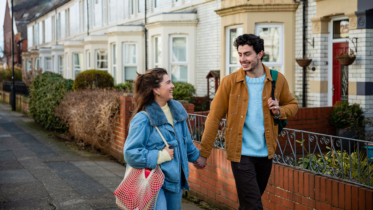 Couple walking hand-in-hand along a residential street with brick houses and gardens in the background.