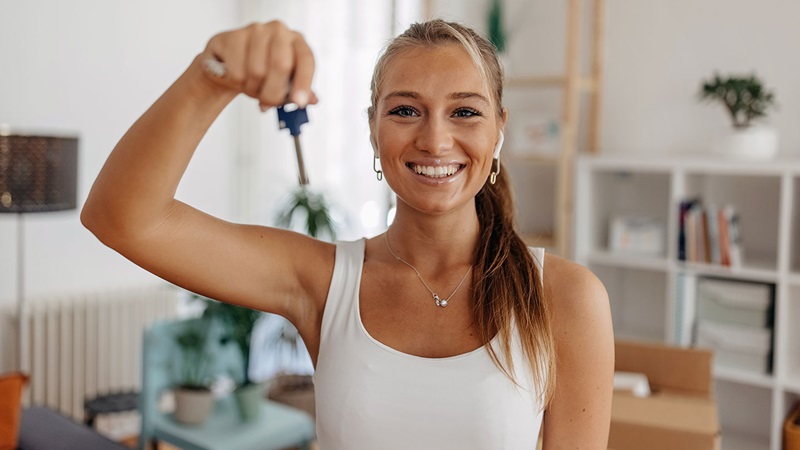 Happy young woman holding up a key in a bright, modern home interior with boxes and furniture in the background, symbolising moving or new home ownership.