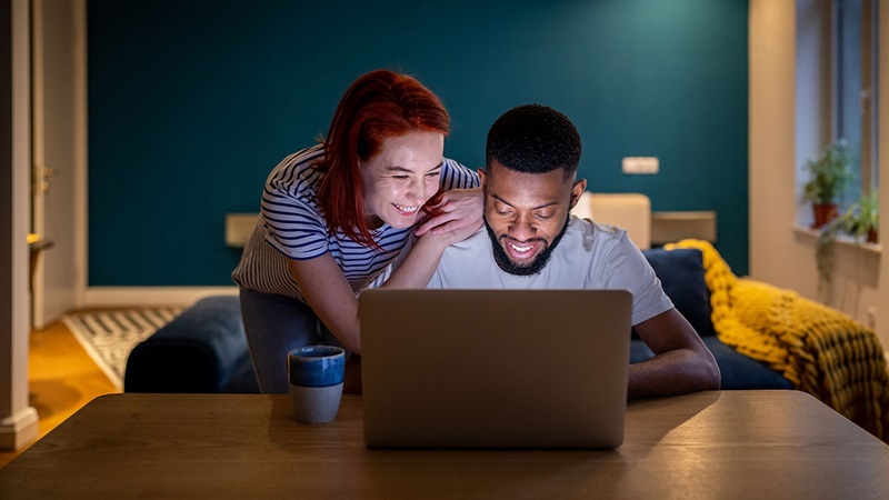 Couple in a cosy living room working on a laptop at a wooden table with a mug nearby, illuminated by warm lighting.