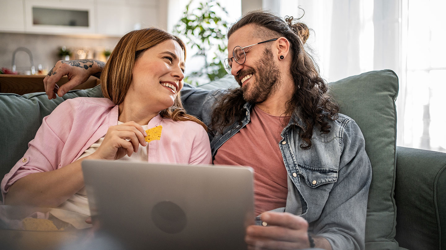 Happy couple sitting on sofa with a laptop smiling at each other whilst eating tortila chips