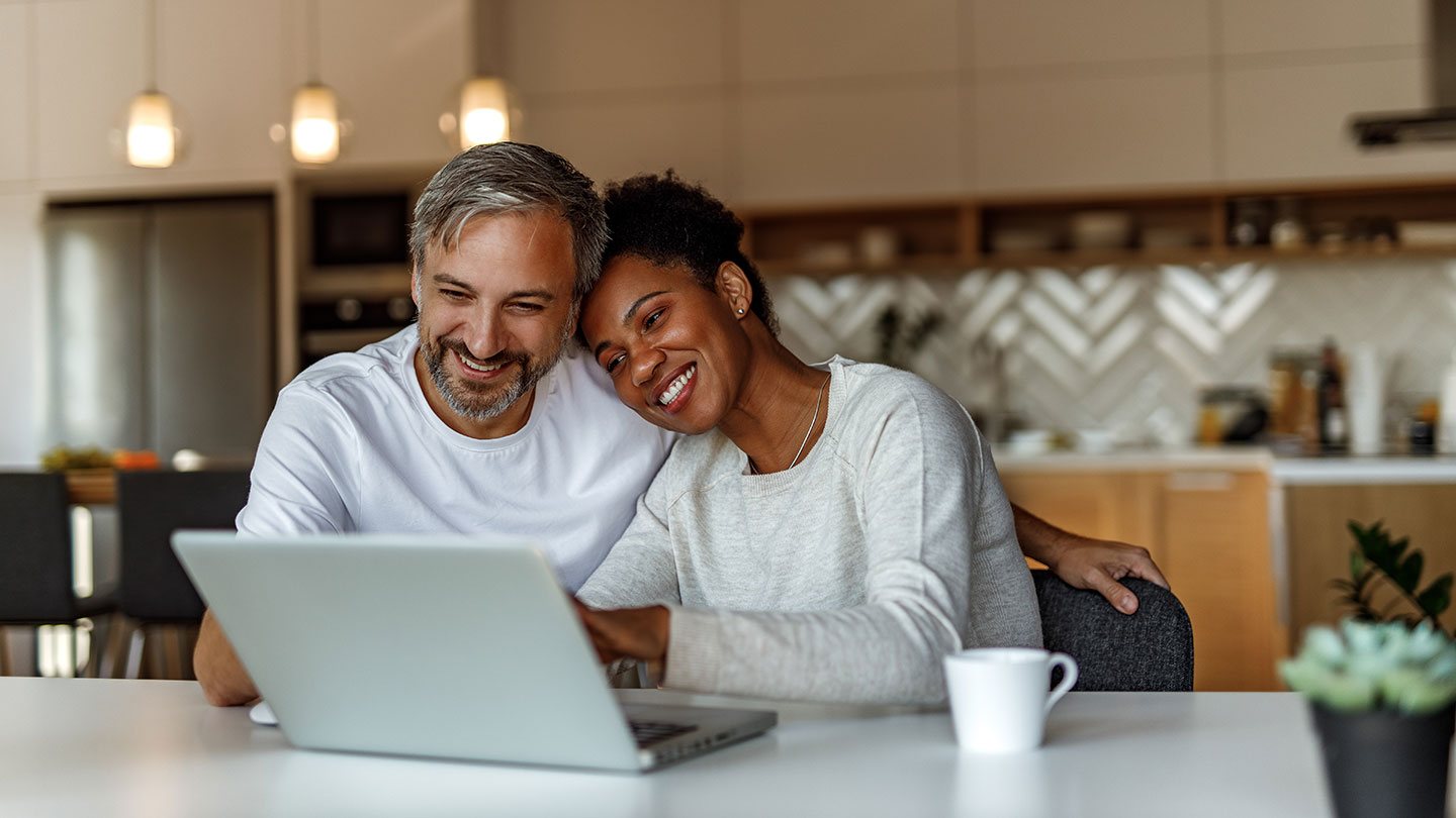 A couple sitting together in their modern kitchen, browsing on a laptop with a mug on the table