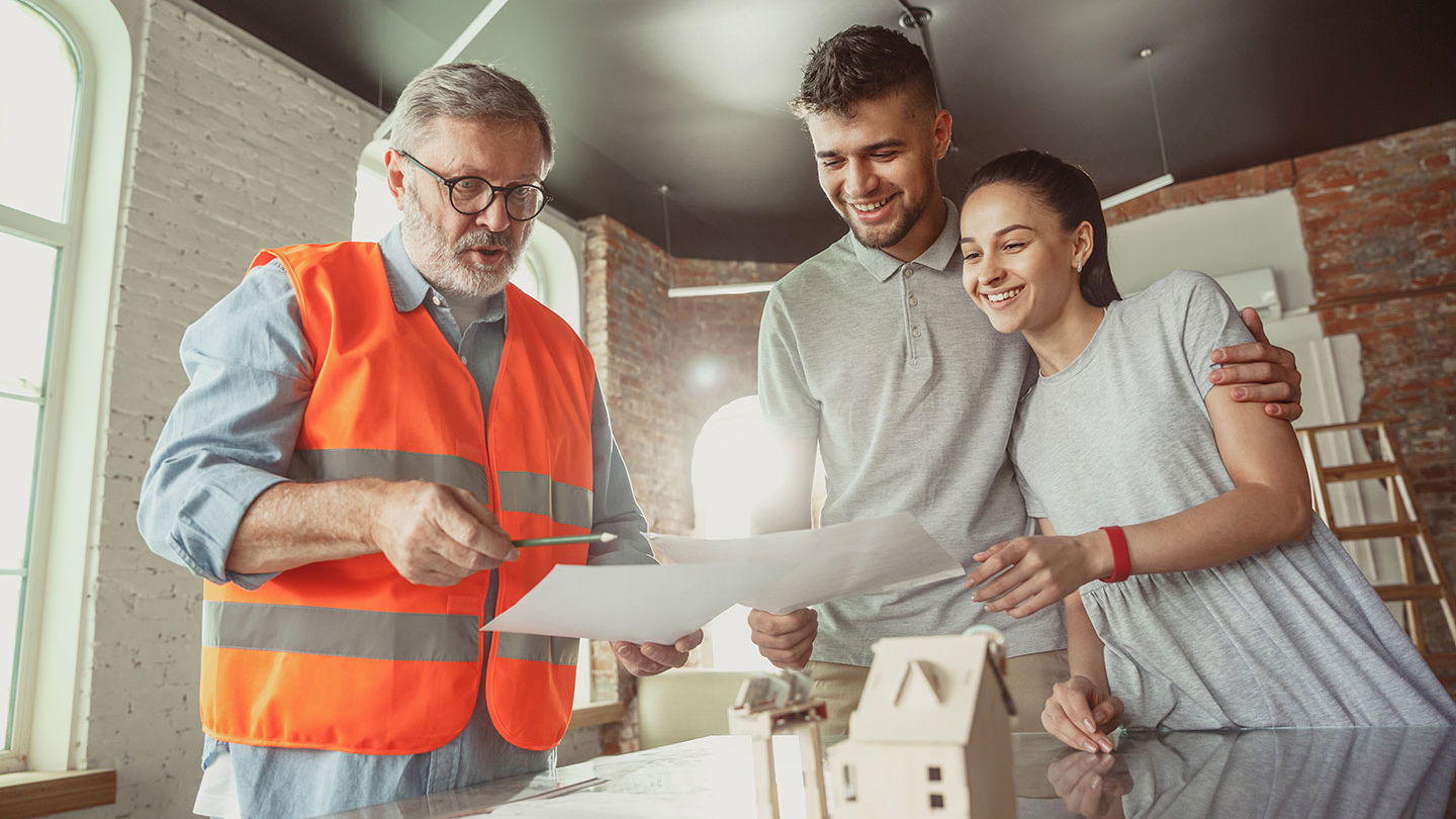 Architect in an orange safety vest discussing building plans with two individuals in a modern interior setting, with a small house model on the table.