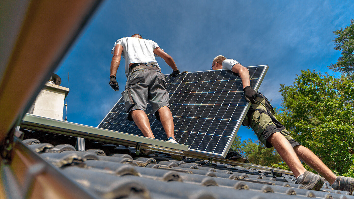 Two men installing a solar panel on a roof, with one man holding the panel while the other assists, under a clear blue sky.