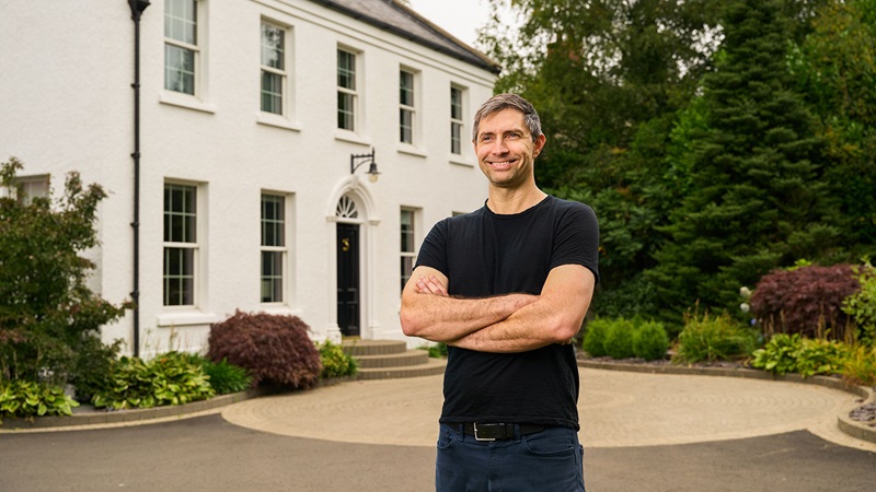 A Danske Bank mortgage customer standing confidently in front of a new, elegant white home with a landscaped driveway and garden.