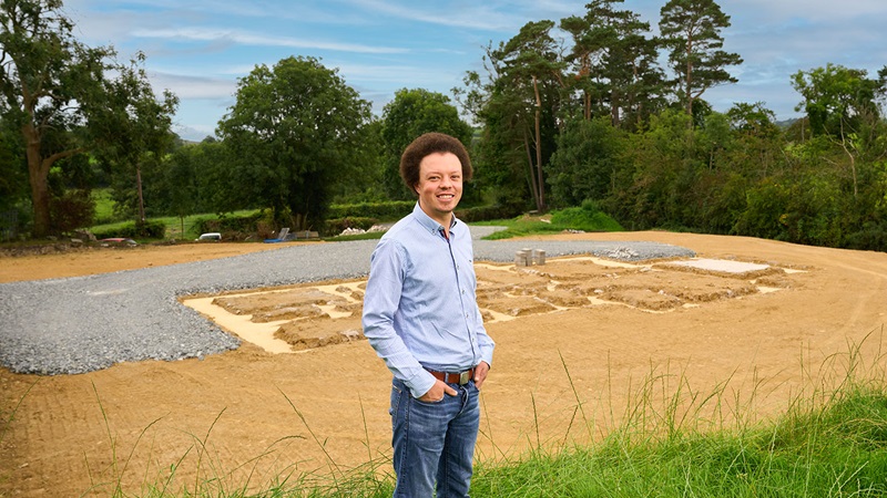 A Danske Bank self-build customer standing at the site of their new home under construction, surrounded by a rural landscape with trees and greenery.