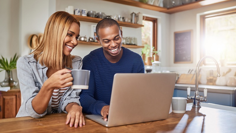 Couple smiling in kitchen whilst using laptop Couple smiling in kitchen whilst using laptop