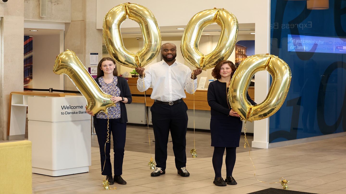 Three people in a bank branch holding balloons that spell out 1000
