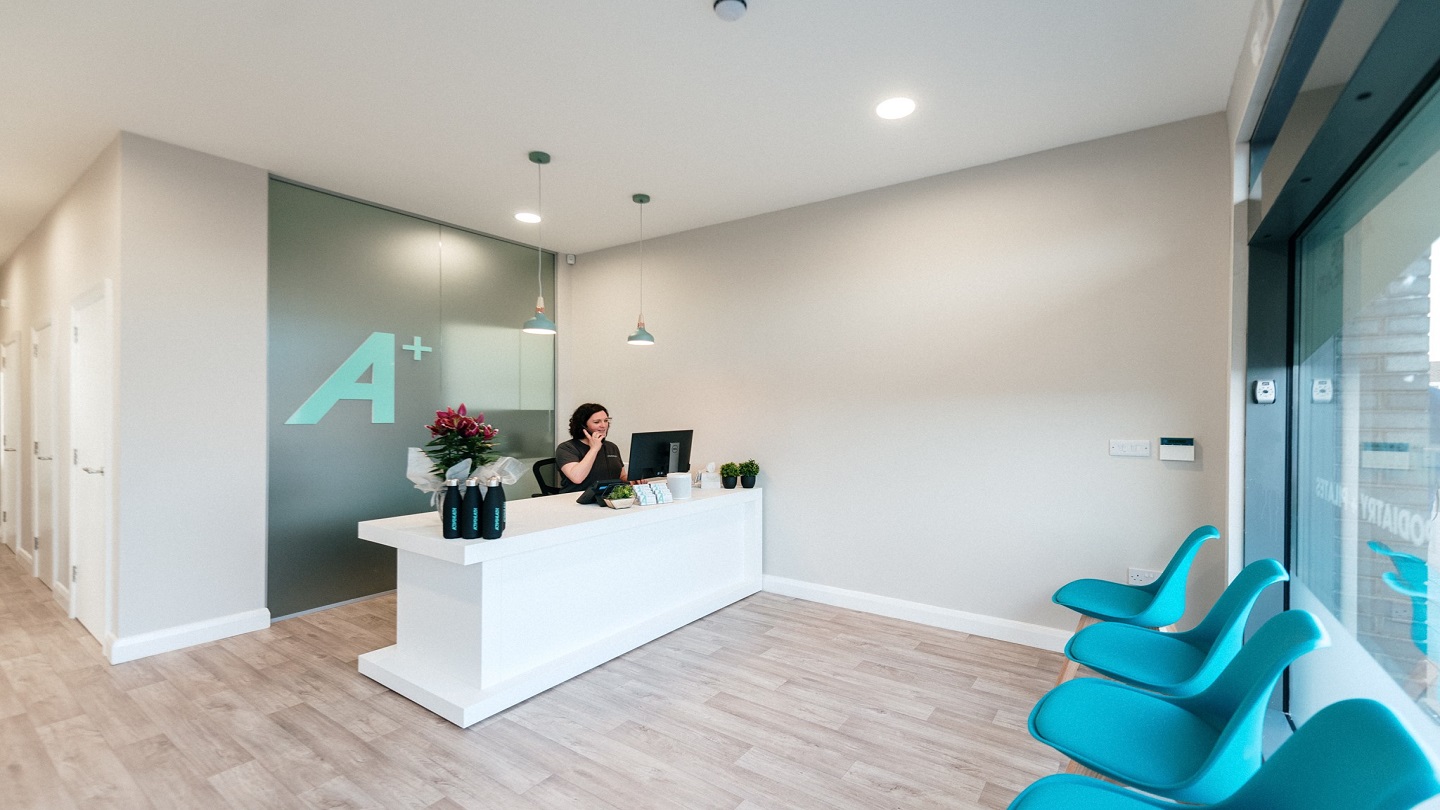 Photo showing an open reception - with a woman at a desk. Colours are white, grey and blue.