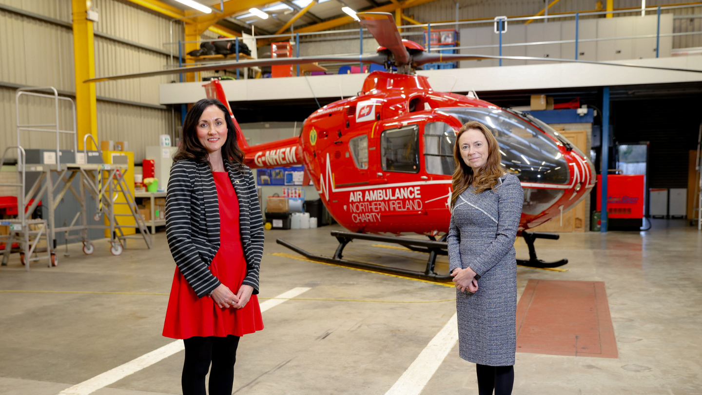 Two women stand inside a large structure. Behind them is the bright red Air Ambulance NI.