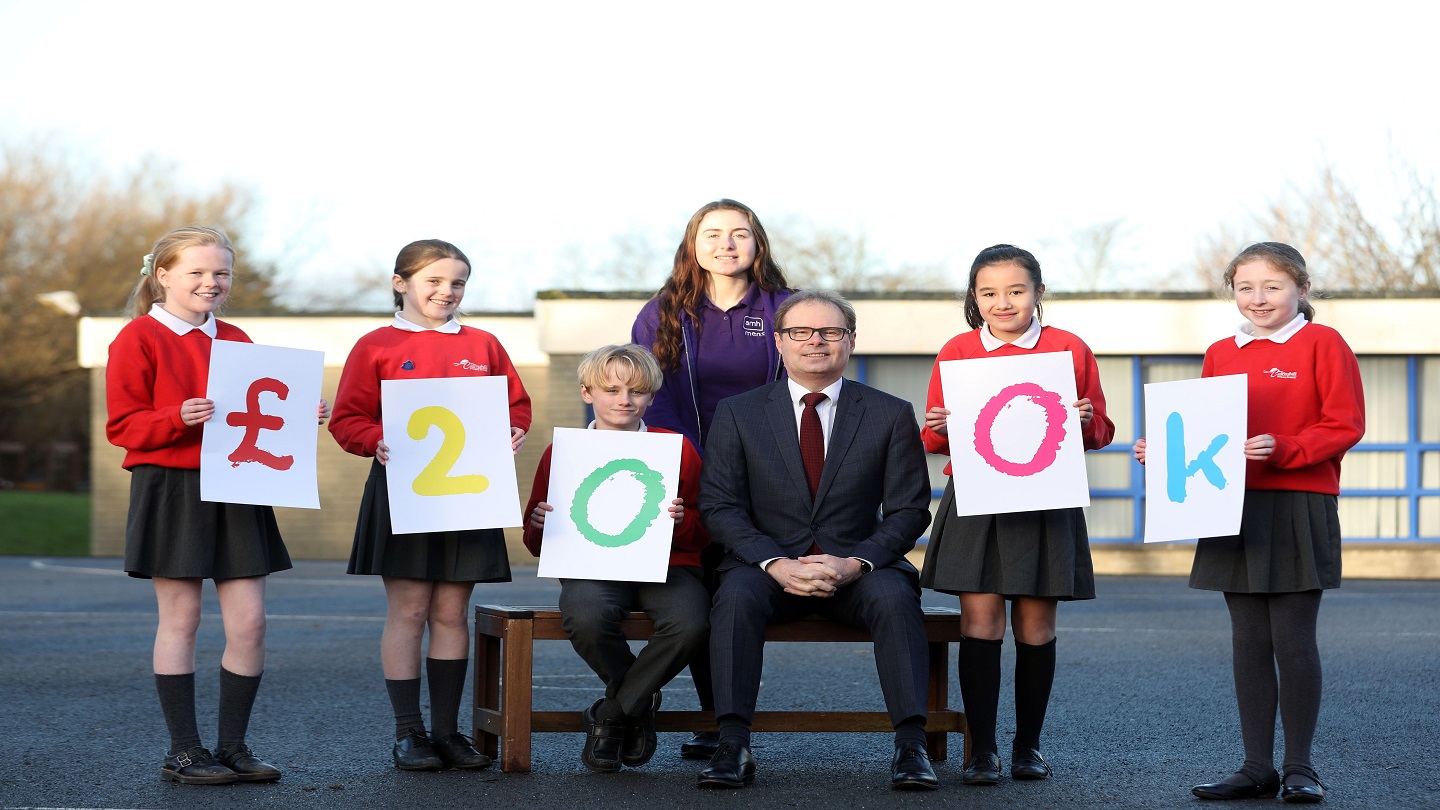 Five school pupils out on a playground. They are wearing red uniforms and holding multicoloured signs that spell out £200k. A man in business attire and a woman in a purple fleece join them in the photo.