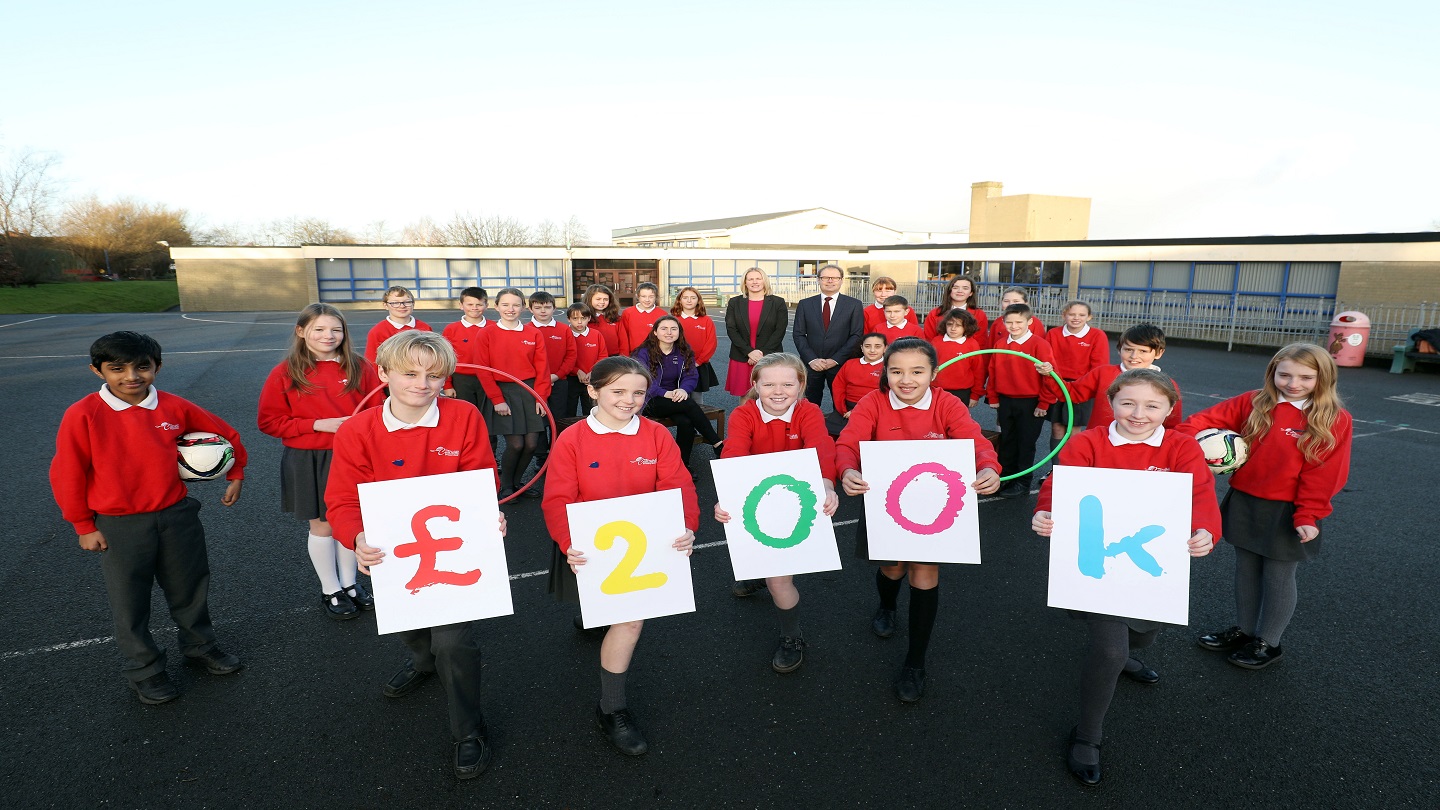 A class of school pupils in red uniforms stand outside in a playground holding up signs that spell out £200k. A woman and man in business attire and a woman in a purple fleece stand with them.