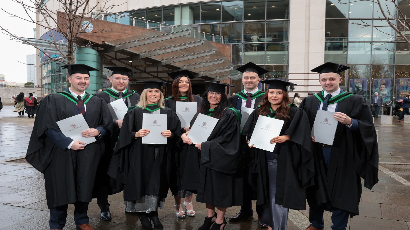 Eight people in graduation robes holding certificates of their degrees.