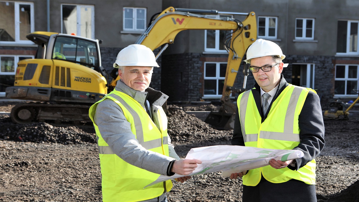 Two men in high vis vests on a housing building site. They're holding a plan and there's a yellow digger behind them.