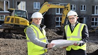 Two men in high vis vests on a housing building site. They're holding a plan and there's a yellow digger behind them.