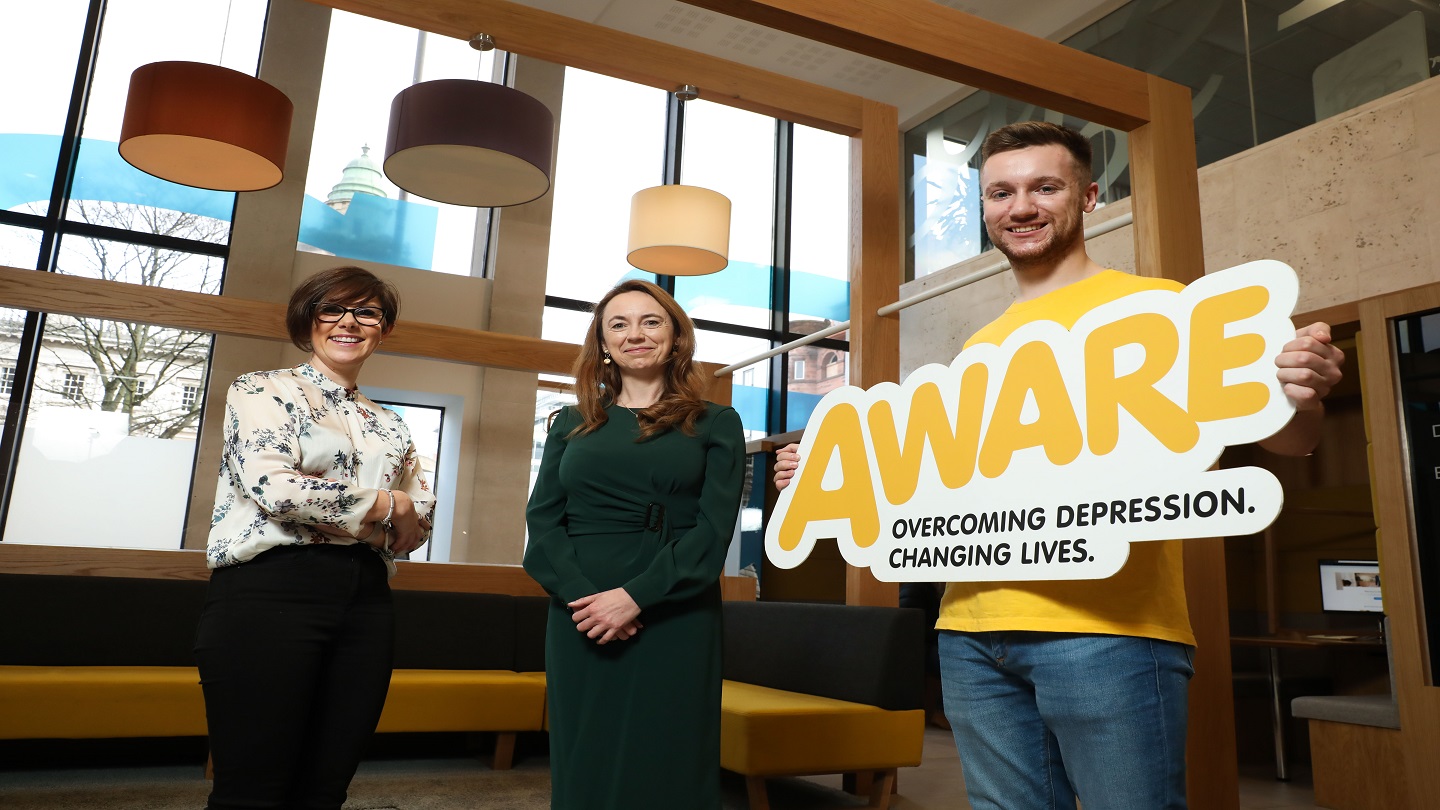 Three people standing inside a Danske Bank branch smiling at the camera. One is holding a large sign with the yellow AWARE logo on it.