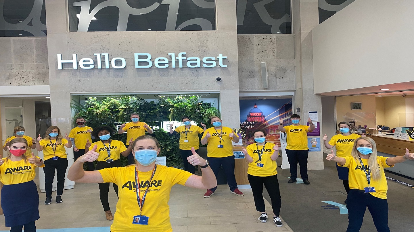 A Danske Bank branch team of 13 people stand socially distanced and with masks on. They have yellow AWARE t shirts on and all have their thumbs up! The 'Hello Belfast' light up sign is in the background.