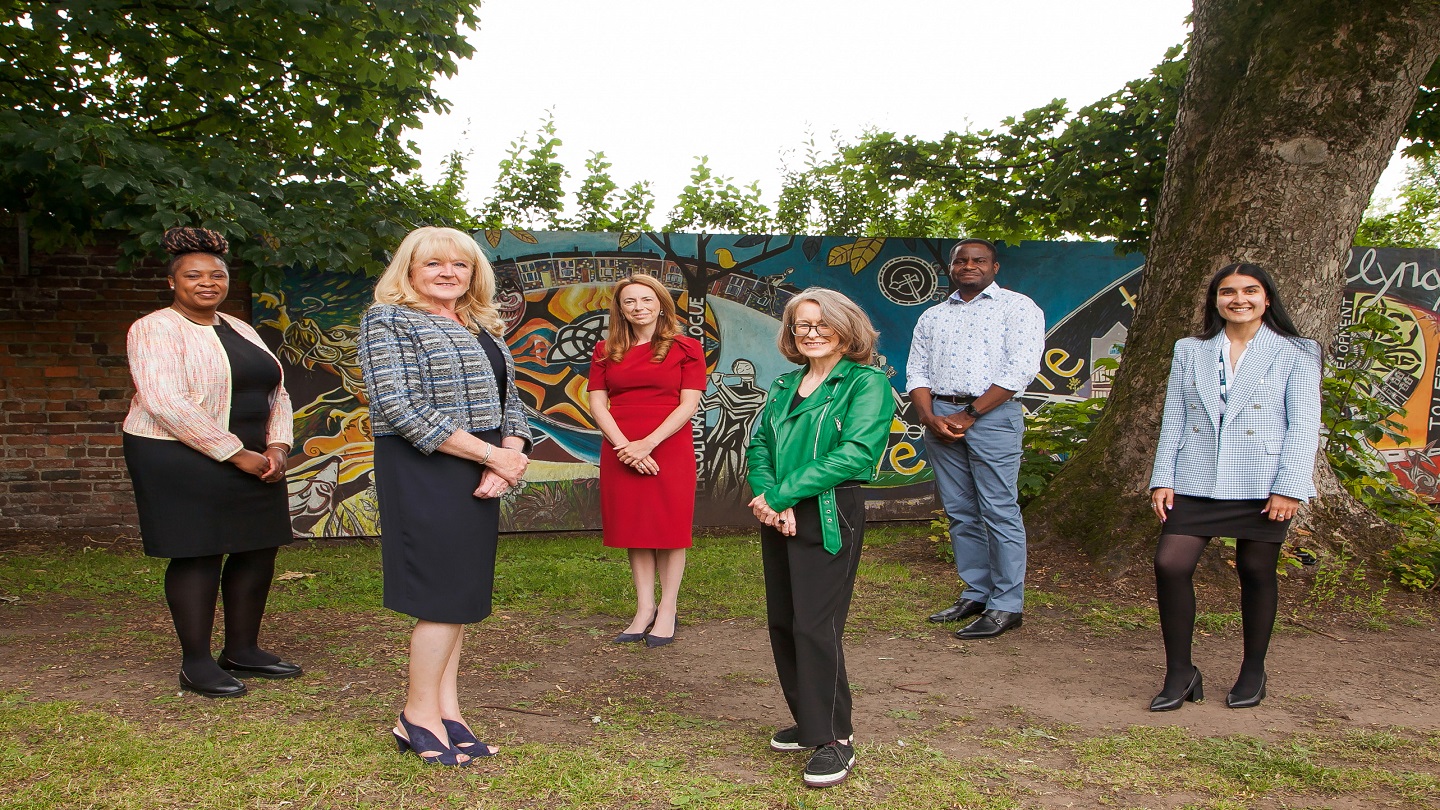 Six people standing outside socially distanced from each other, all looking into the camera and smiling.