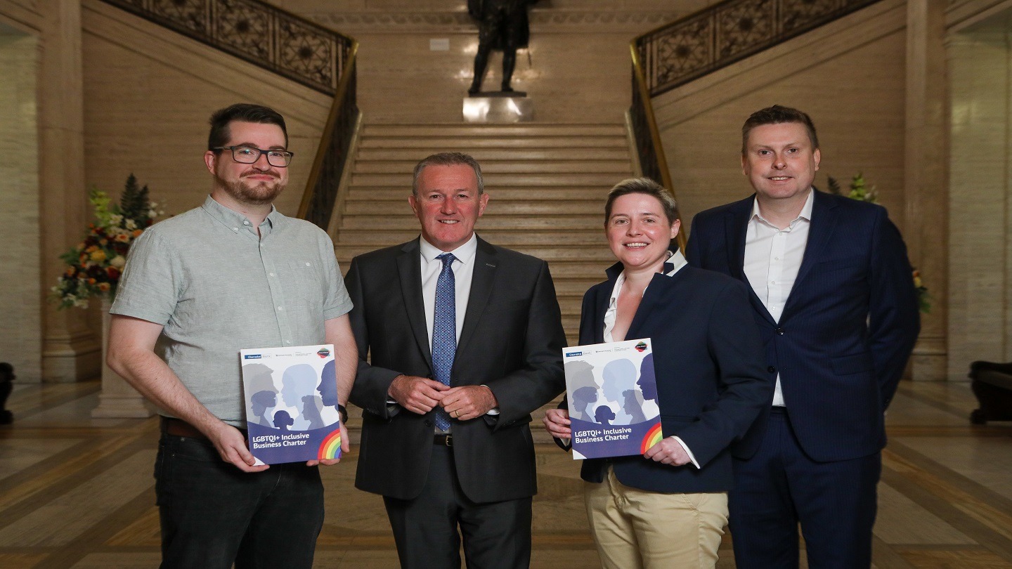 Four people stand together and look into the camera. Two are holding booklets with information on the Cara-Friend LGBTQI+ Inclusive Business Charter.