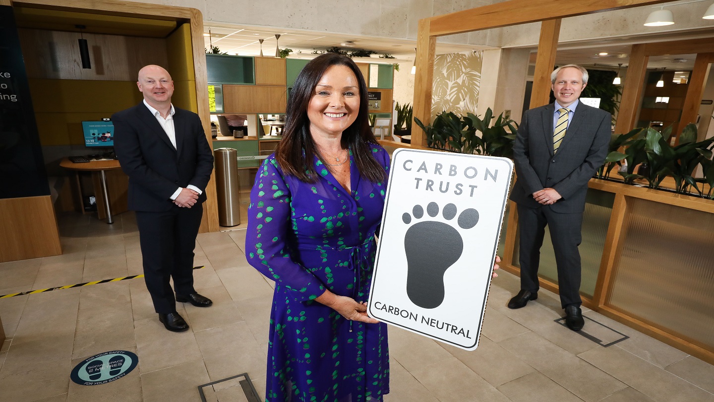 Three people pictured standing socially distanced from each other in a bright Danske Bank branch. At the front, a person is holding a cut out of the Carbon trust logo, which is a black footprint and the words Carbon Trust at the top.