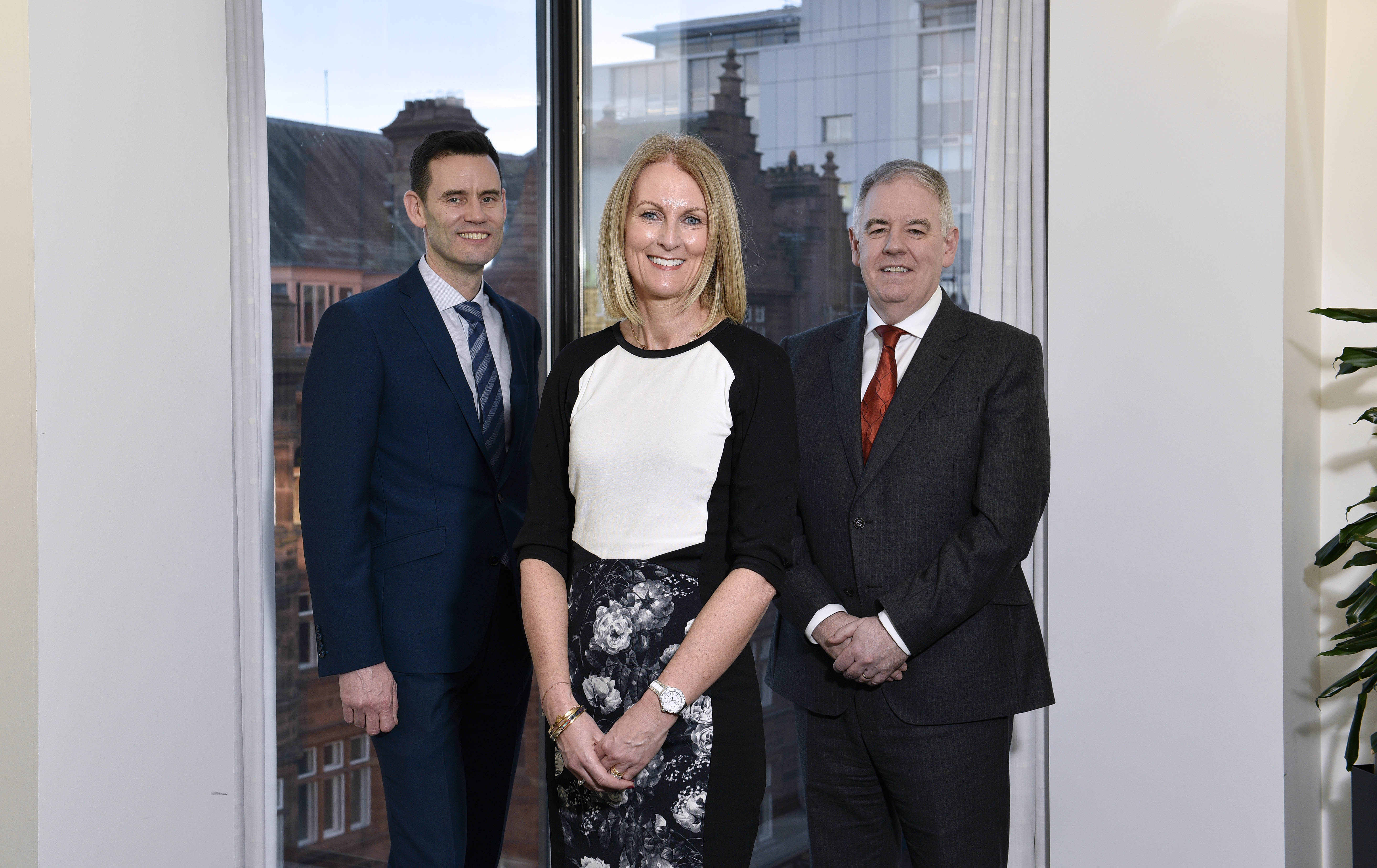 Two men in suits stand in front of a window, a woman stands in front of them in the middle, smiling.