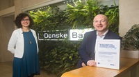 A woman and man stand distanced from each other inside a Danske Bank branch. Behind them is a green living wall with lots of leaves, and a white light box with the Danske Bank logo. Both people are smiling, and looking at the camer. The man is on the right and is leaning on a counter, and holding a framed certificate.