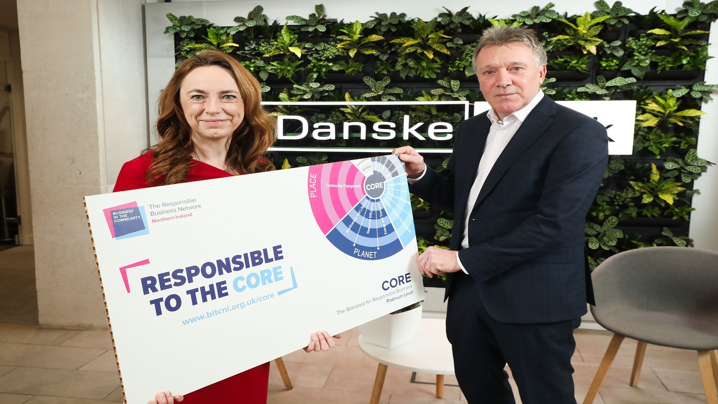 Two business people standing inside a Danske Bank branch. They are holding a Business in the Community 'Responsible to the CORE' sign.