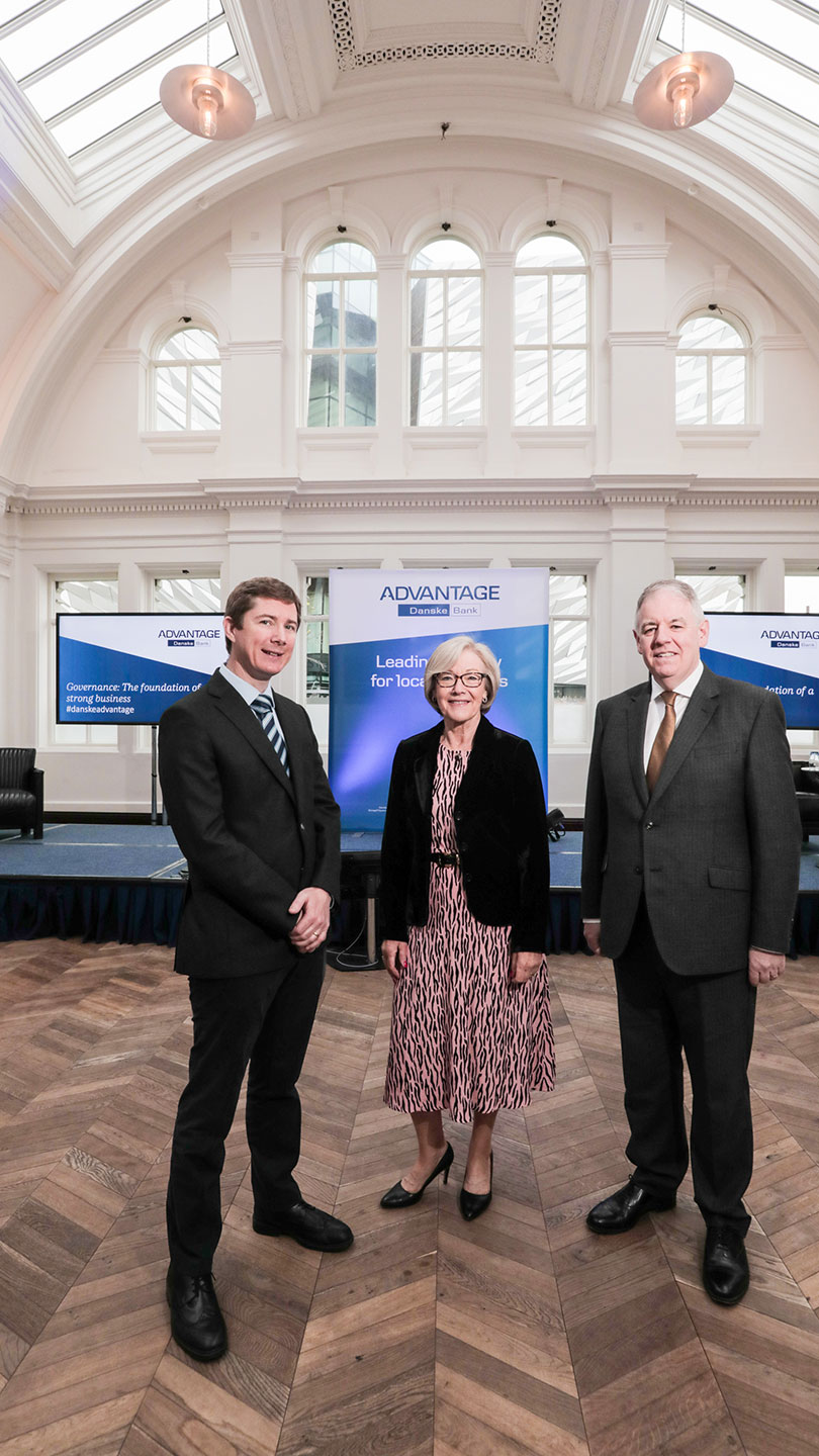 Three people stand in a big bright room, all dressed very smartly. Behind them, on screens and a pop up, is the navy and white Danske Advantage branding.