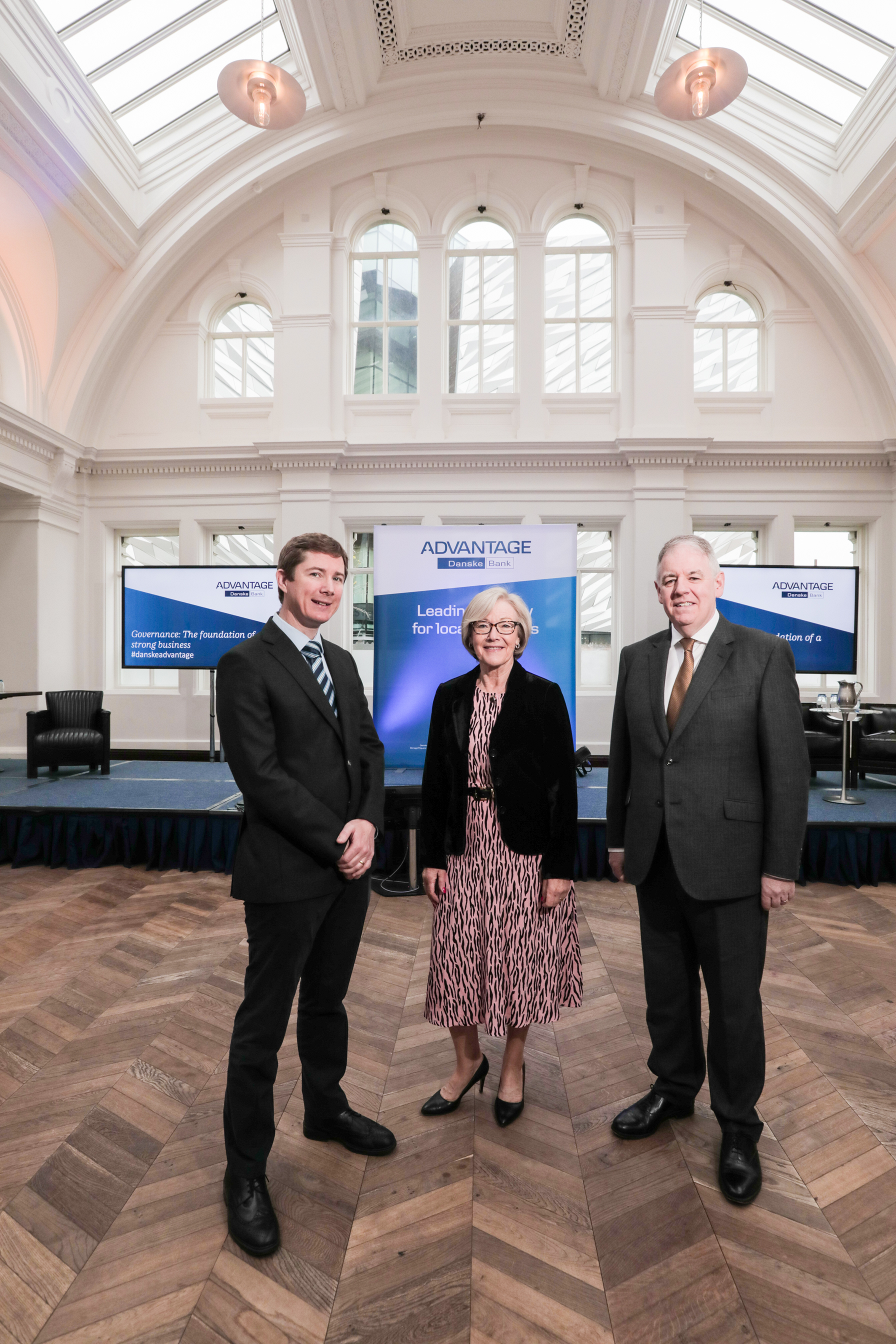 Three people stand in a big bright room, all dressed very smartly. Behind them, on screens and a pop up, is the navy and white Danske Advantage branding.