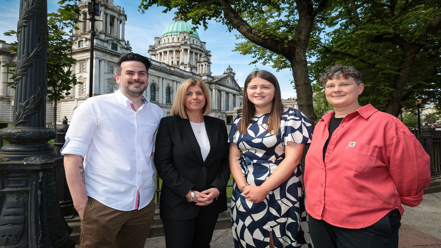 Four people stand outside, with Belfast City Hall in the background. They're all smiling at the camera.